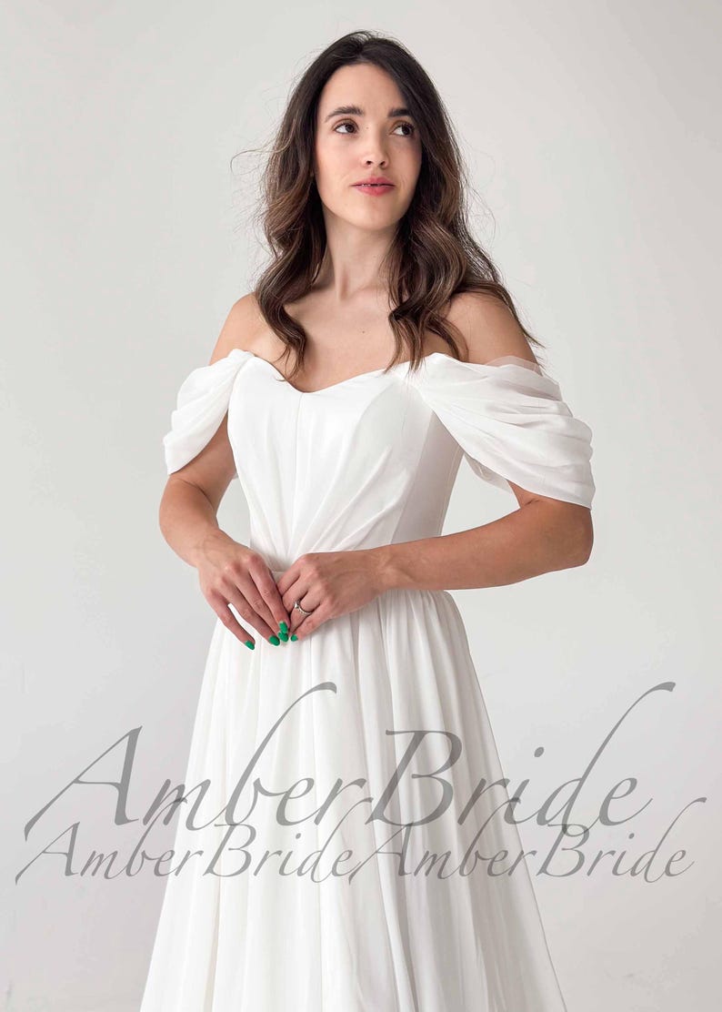 May include: A white off-the-shoulder wedding dress with a flowing skirt. The dress features draped sleeves and a sweetheart neckline. The text "Amber Bride" is visible in a script font at the bottom of the image.
