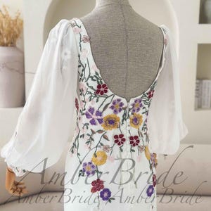 May include: White wedding dress with floral embroidery in purple, yellow, and red. The dress features sheer, long sleeves and a low back. The dress is displayed on a mannequin.