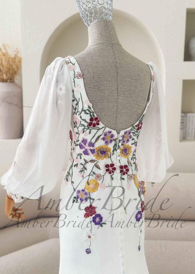 May include: White wedding dress with floral embroidery in purple, yellow, and red. The dress features sheer, long sleeves and a low back. The dress is displayed on a mannequin.