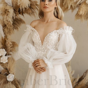 May include: A white off-the-shoulder wedding dress with a plunging neckline and long, puffy sleeves. The dress has a fitted bodice and a flowing skirt. The model is standing in front of a backdrop of dried pampas grass.