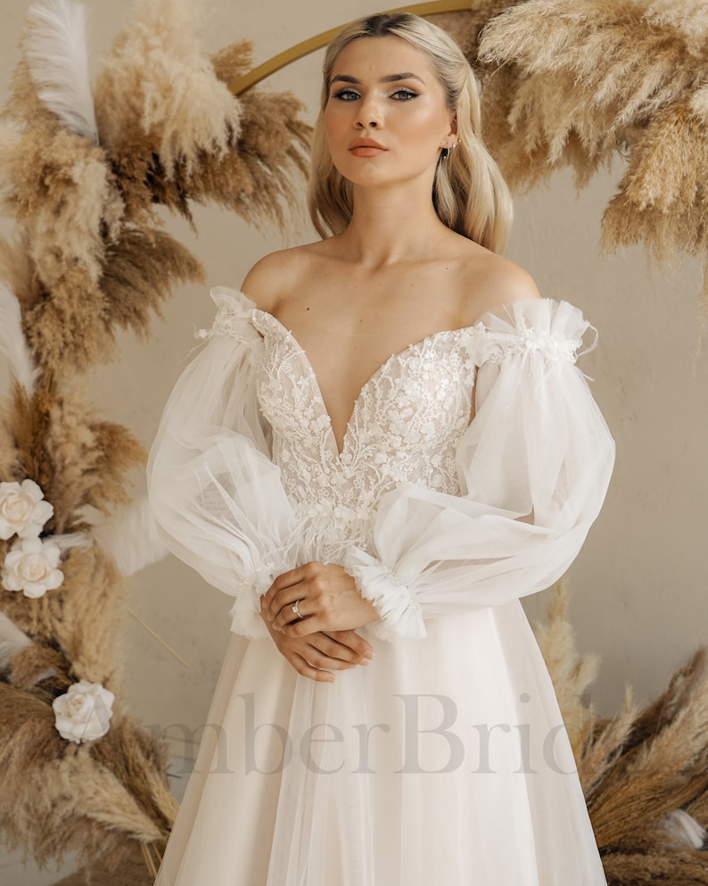 May include: A white off-the-shoulder wedding dress with a plunging neckline and long, puffy sleeves. The dress has a fitted bodice and a flowing skirt. The model is standing in front of a backdrop of dried pampas grass.