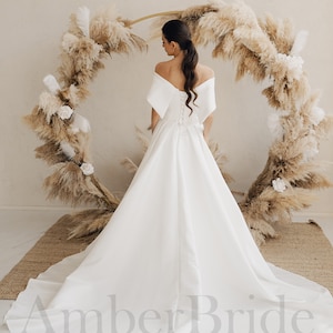 May include: A white off-the-shoulder wedding dress with a fitted bodice and a flowing skirt. The dress has a lace-up back and a train. The dress is photographed against a backdrop of dried pampas grass.