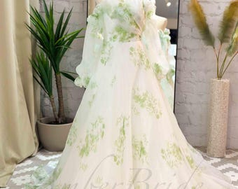 May include: A floor-length off-the-shoulder gown featuring sheer white fabric with a light green floral print. The dress has long, voluminous sleeves, a fitted bodice, and a full, flowing skirt with a train. The text AmberBride is visible.