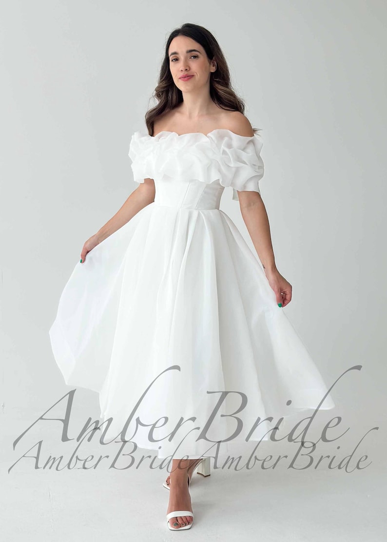 May include: A white off-the-shoulder dress with a ruffled neckline and short sleeves. The dress has a fitted bodice and a flowing skirt. The text "Amber Bride" is visible at the bottom of the image. The dress is suitable for a wedding or special occasion.