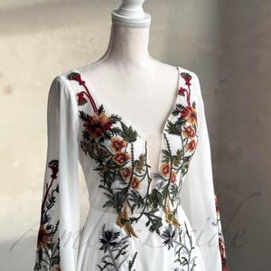 May include: A white maxi dress with long sleeves and a deep V-neckline. The dress is adorned with colorful floral embroidery in shades of red, yellow, green, and brown.