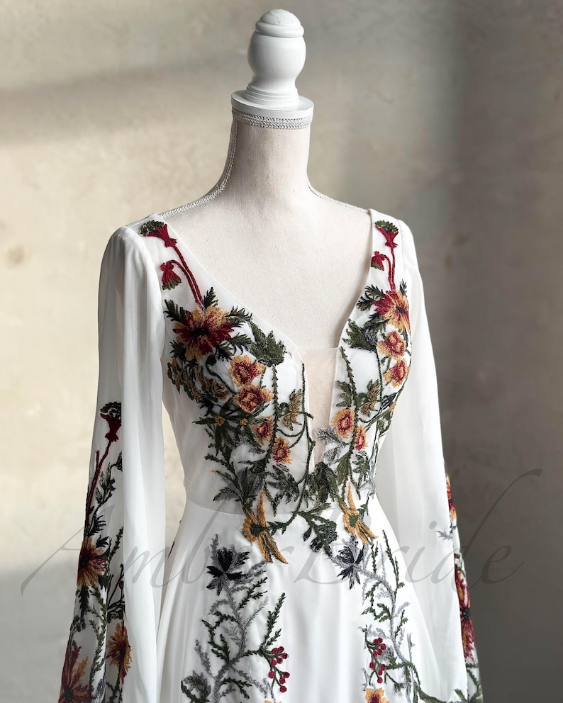 May include: A white maxi dress with long sleeves and a deep V-neckline. The dress is adorned with colorful floral embroidery in shades of red, yellow, green, and brown.