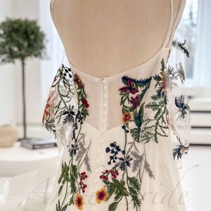 May include: A white wedding dress with a floral embroidery design. The dress has a fitted bodice and a flowing skirt. The embroidery features a variety of flowers and leaves in shades of red, green, blue, and yellow.