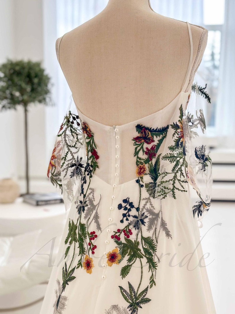 May include: A white wedding dress with a floral embroidery design. The dress has a fitted bodice and a flowing skirt. The embroidery features a variety of flowers and leaves in shades of red, green, blue, and yellow.