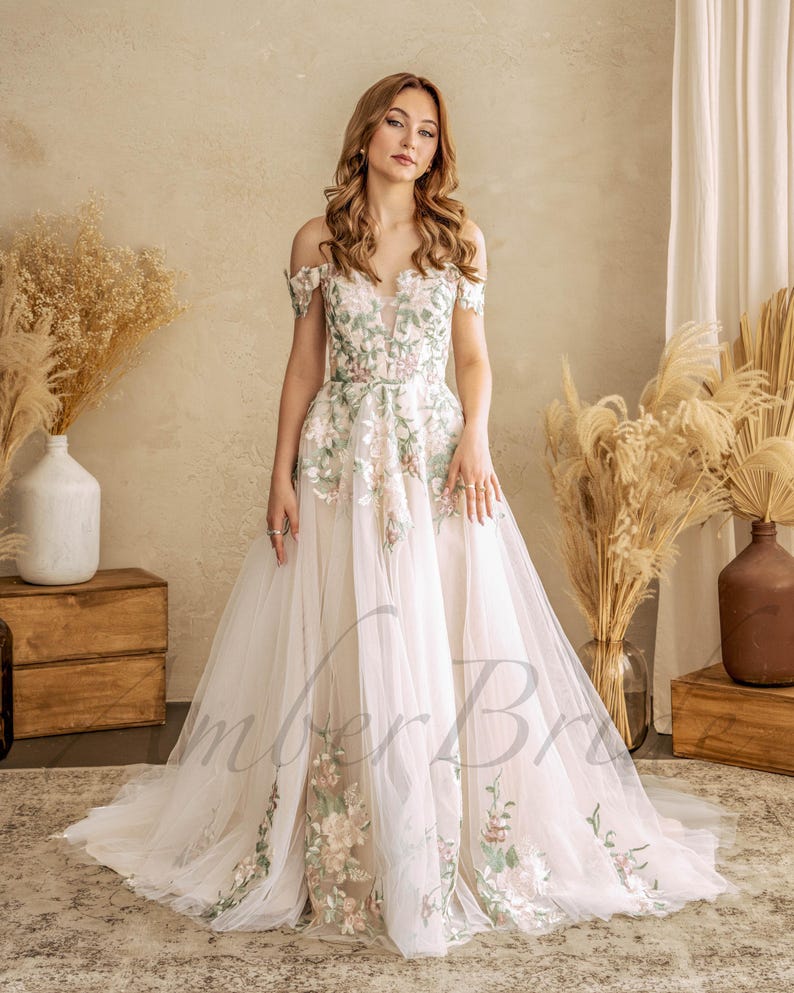 May include: A white off-the-shoulder wedding dress with floral embroidery in shades of green, pink, and white. The dress has a fitted bodice and a flowing skirt.
