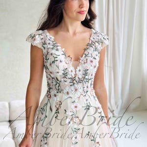 May include: A cream-colored wedding dress with a V-neckline and cap sleeves. The dress is adorned with delicate floral embroidery in white, pink, and green. The skirt flows gracefully, creating an elegant silhouette. The text "Amber Bride" is visible.