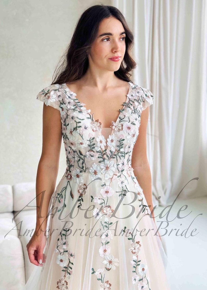 May include: A cream-colored wedding dress with a V-neckline and cap sleeves. The dress is adorned with delicate floral embroidery in white, pink, and green. The skirt flows gracefully, creating an elegant silhouette. The text "Amber Bride" is visible.