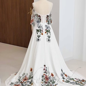 May include: A white maxi dress with a floral embroidery design. The dress has a scoop neckline and thin straps. The embroidery features a variety of colorful flowers and leaves.