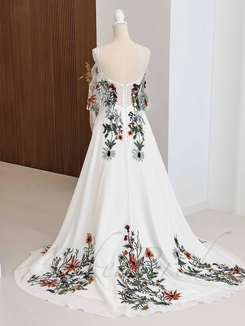 May include: A white maxi dress with a floral embroidery design. The dress has a scoop neckline and thin straps. The embroidery features a variety of colorful flowers and leaves.