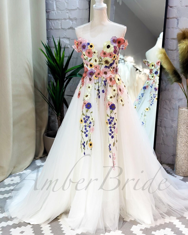 May include: A white off-the-shoulder tulle gown with colorful floral embroidery. The dress has a fitted bodice and a flowing skirt. The embroidery features pink, yellow, blue, and green flowers.