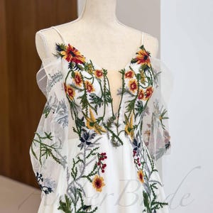 May include: A white wedding dress with a floral embroidery design. The dress has a V-neckline and a flowing skirt. The embroidery features yellow, red, and green flowers.