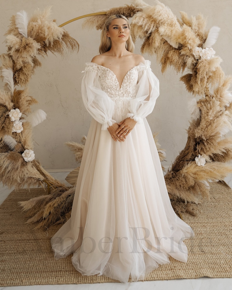 May include: A white off-the-shoulder wedding dress with long, puffy sleeves and a fitted bodice. The dress has a flowing skirt and is made of a delicate fabric.