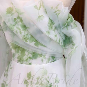 May include: A white tulle dress with a green floral print. The dress has a one-shoulder design and a flowing skirt.