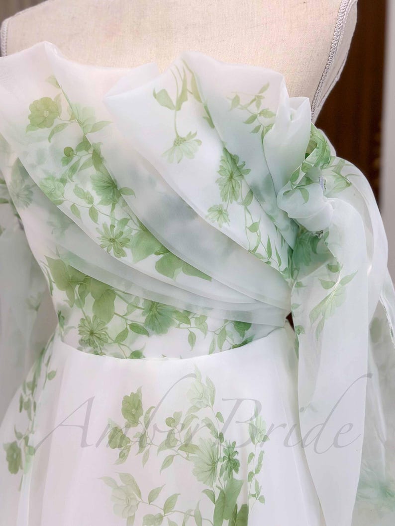 May include: A white tulle dress with a green floral print. The dress has a one-shoulder design and a flowing skirt.