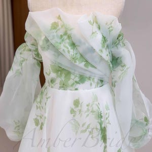 May include: A white off-the-shoulder gown with a floral print in shades of green. The gown has a ruffled neckline and long, puffy sleeves. The skirt is full and flowing.