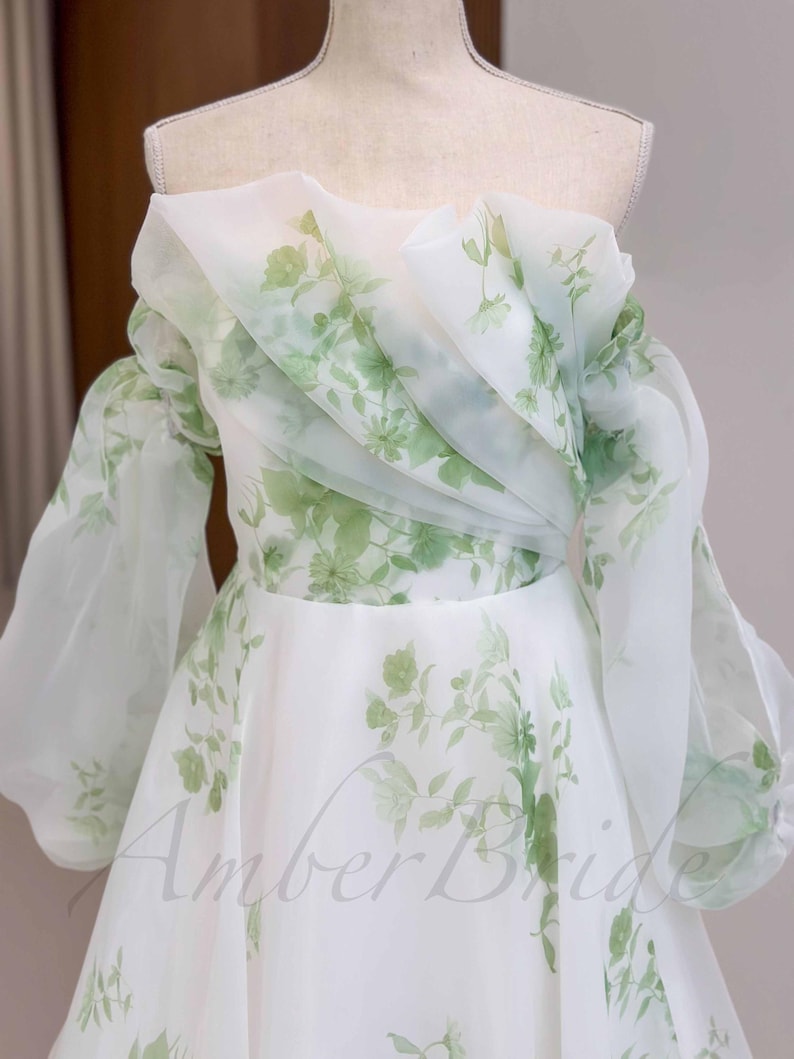 May include: A white off-the-shoulder gown with a floral print in shades of green. The gown has a ruffled neckline and long, puffy sleeves. The skirt is full and flowing.