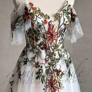 May include: White tulle dress with embroidered floral details in shades of green, red, and yellow. The dress has a V-neckline and short, ruffled sleeves.