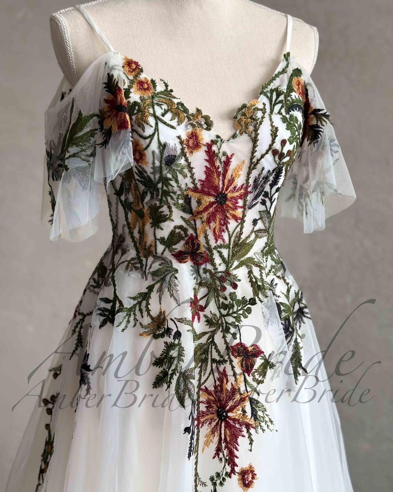 May include: White tulle dress with embroidered floral details in shades of green, red, and yellow. The dress has a V-neckline and short, ruffled sleeves.