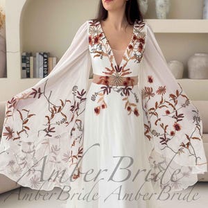 May include: A white wedding dress with a V-neckline and long, flowing sleeves. The dress features floral embroidery in shades of brown and red. The text "Amber Bride" is visible on the bottom of the dress.