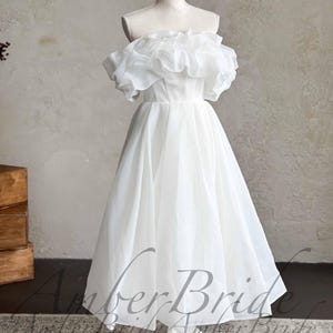 May include: A white, off-the-shoulder wedding dress with a ruffled neckline and a tea-length skirt. The dress is displayed on a mannequin. The dress has a simple, elegant design, perfect for a wedding or special occasion.