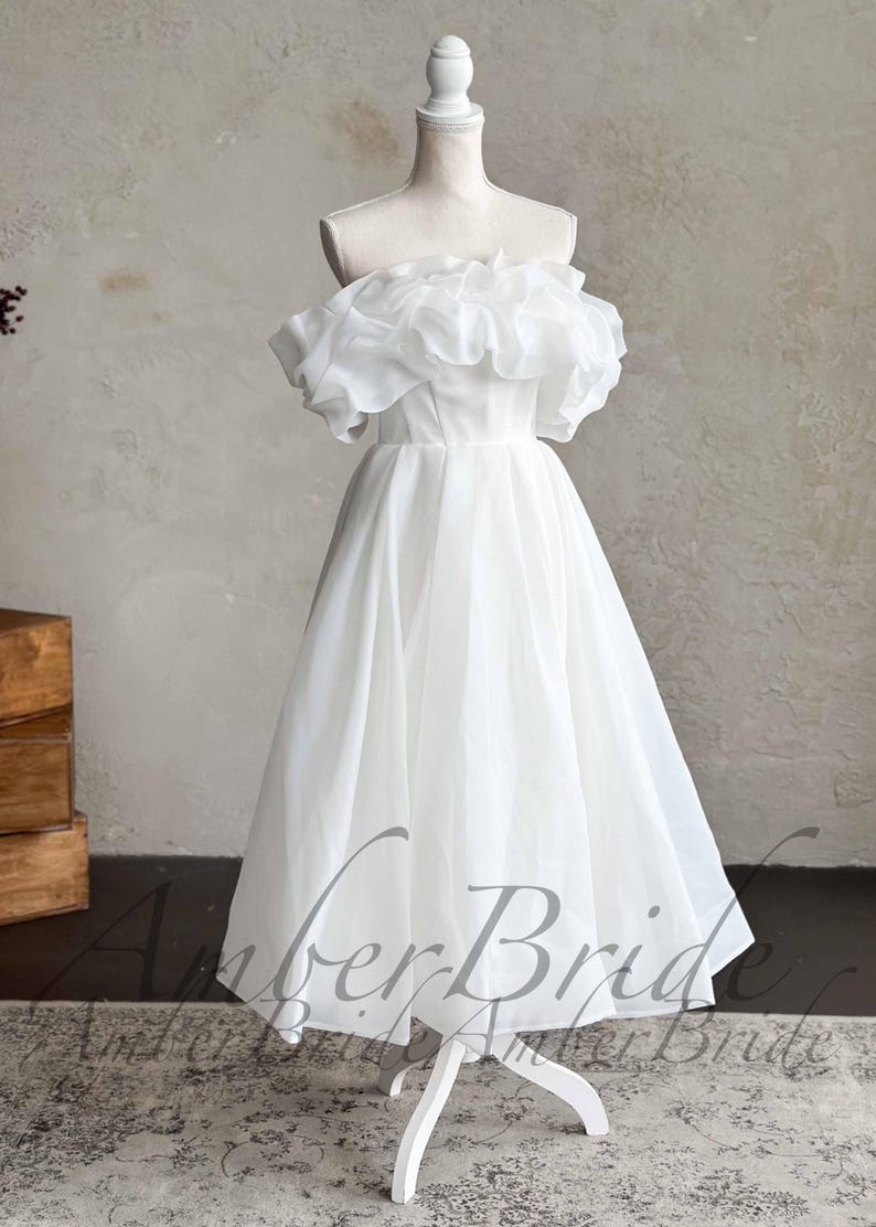 May include: A white, off-the-shoulder wedding dress with a ruffled neckline and a tea-length skirt. The dress is displayed on a mannequin. The dress has a simple, elegant design, perfect for a wedding or special occasion.