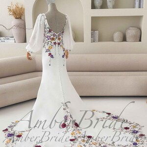 May include: White wedding dress with floral embroidery. The dress features a low back, long sheer sleeves, and a long train with floral detailing. The text "Amber Bride" is visible on the train.