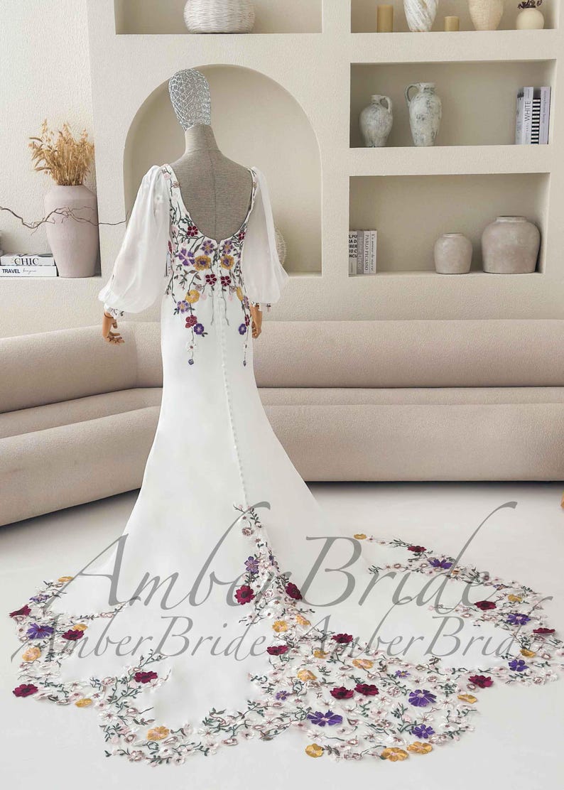 May include: White wedding dress with floral embroidery. The dress features a low back, long sheer sleeves, and a long train with floral detailing. The text "Amber Bride" is visible on the train.