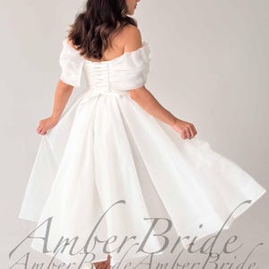 May include: A white off-the-shoulder wedding dress with short, puffy sleeves and a tea-length skirt. The dress features a corset back and is made of a flowing, lightweight fabric. The text "Amber Bride" is visible.