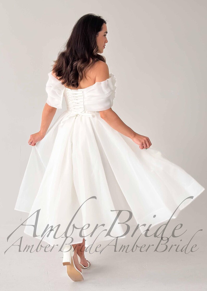 May include: A white off-the-shoulder wedding dress with short, puffy sleeves and a tea-length skirt. The dress features a corset back and is made of a flowing, lightweight fabric. The text "Amber Bride" is visible.