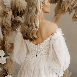 May include: A white off-the-shoulder wedding dress with long, puffy sleeves and a lace bodice. The dress has a tulle skirt and a train. The back of the dress features a row of buttons and a feather trim.