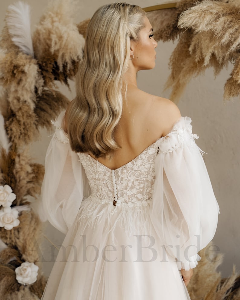 May include: A white off-the-shoulder wedding dress with long, puffy sleeves and a lace bodice. The dress has a tulle skirt and a train. The back of the dress features a row of buttons and a feather trim.