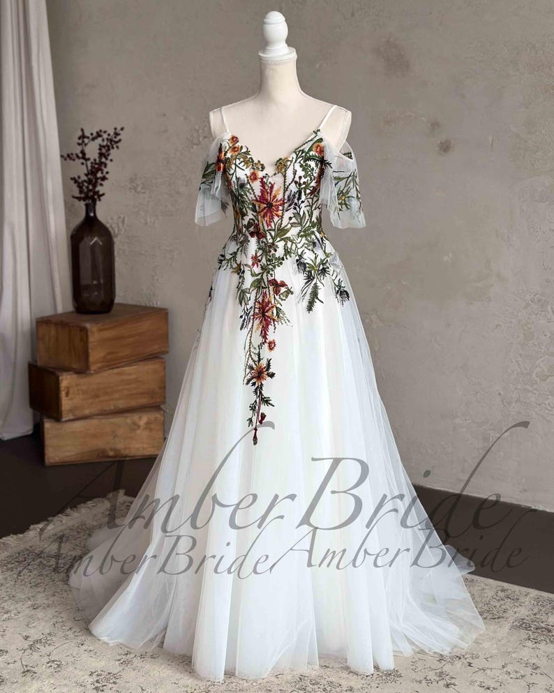 May include: A white tulle gown with a floral embroidery design. The dress has a V-neckline, off-the-shoulder straps, and a flowing skirt. The embroidery features a variety of flowers in shades of red, orange, yellow, and green.