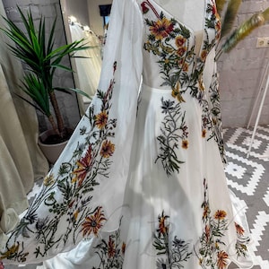 May include: A white maxi dress with long sleeves and a deep V-neckline. The dress is covered in a floral pattern with red, yellow, and green flowers.