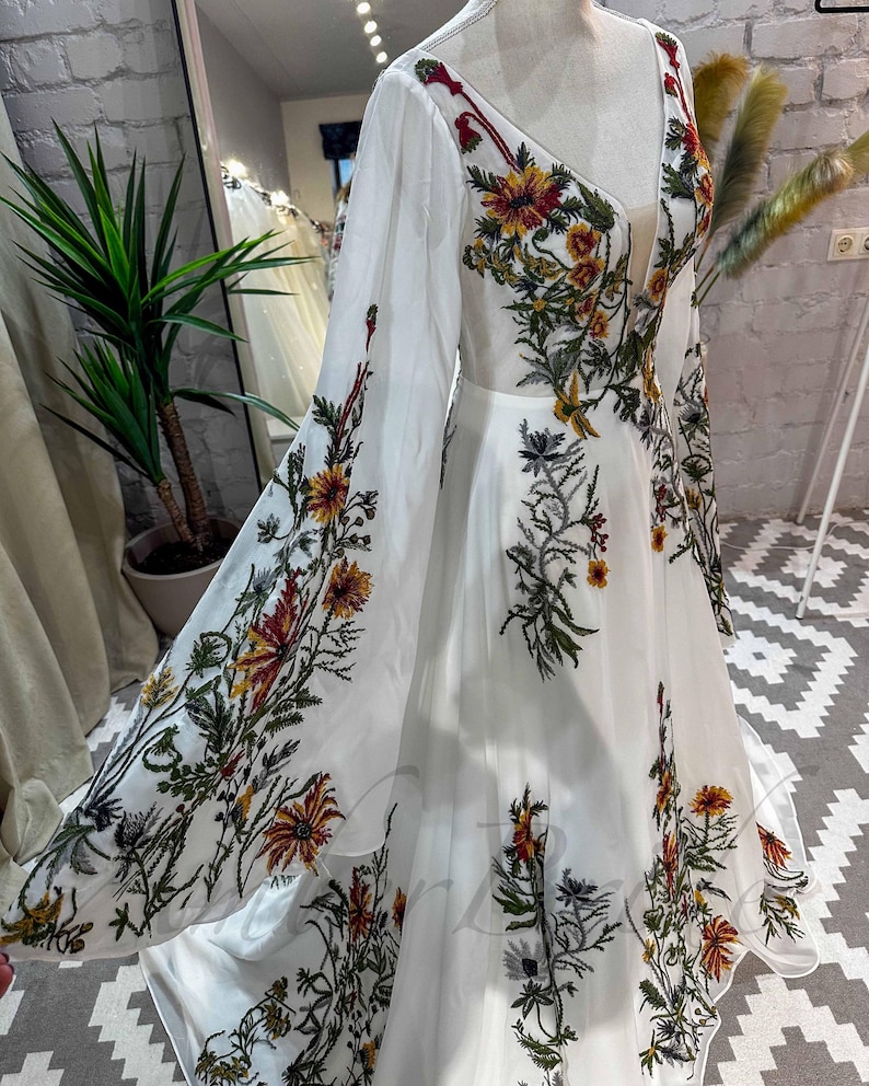 May include: A white maxi dress with long sleeves and a deep V-neckline. The dress is covered in a floral pattern with red, yellow, and green flowers.