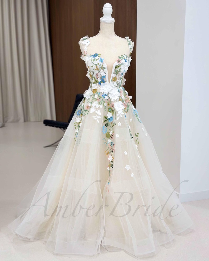 May include: A white tulle gown with a plunging neckline and floral embroidery. The embroidery features white, pink, blue, and green flowers.