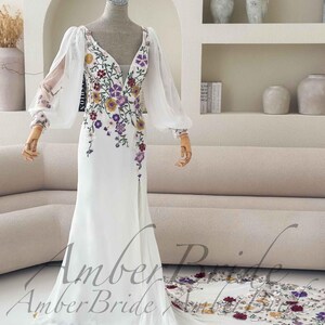 May include: A white wedding dress with a V-neckline and long, sheer sleeves. The dress is adorned with colorful floral embroidery and has a flowing train. The text "Amber Bride" is visible.