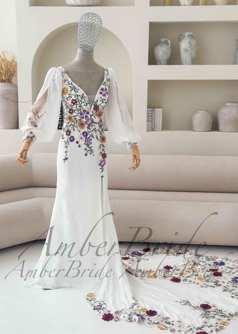May include: A white wedding dress with a V-neckline and long, sheer sleeves. The dress is adorned with colorful floral embroidery and has a flowing train. The text "Amber Bride" is visible.