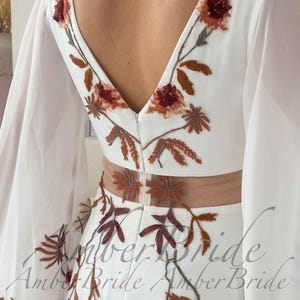 May include: White wedding dress with a V-neckline and sheer sleeves. The dress features floral embroidery in shades of brown and burgundy. The back of the dress has a low V-neck and a sheer panel at the waist. The text "Amber Bride" is visible.