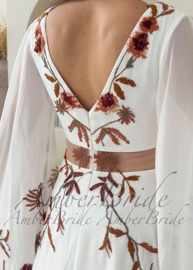 May include: White wedding dress with a V-neckline and sheer sleeves. The dress features floral embroidery in shades of brown and burgundy. The back of the dress has a low V-neck and a sheer panel at the waist. The text "Amber Bride" is visible.