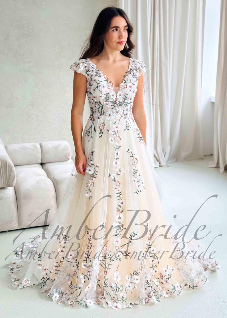 May include: A cream-colored wedding dress with floral embroidery. The dress features a V-neckline, cap sleeves, and a flowing skirt. The embroidery includes white and pink flowers with green leaves. The text "Amber Bride" is visible.