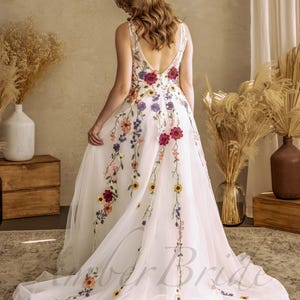 May include: A white tulle wedding dress with a floral embroidery design. The dress has a V-neckline and a low back. The embroidery features a variety of colorful flowers, including red, yellow, blue, and pink.
