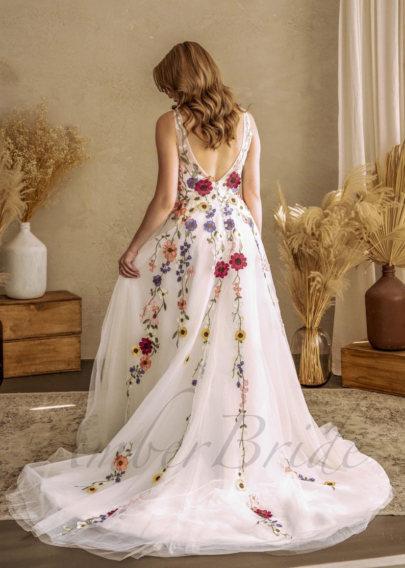 May include: A white tulle wedding dress with a floral embroidery design. The dress has a V-neckline and a low back. The embroidery features a variety of colorful flowers, including red, yellow, blue, and pink.