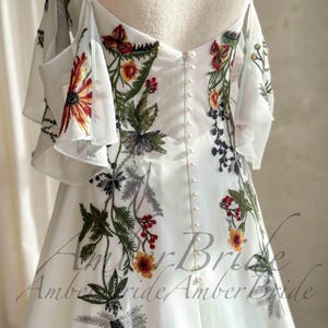 May include: White wedding dress with floral embroidery in red, yellow, green, and black. The dress features a button-down back and off-the-shoulder sleeves with ruffled detailing. The dress has a flowing, elegant design.