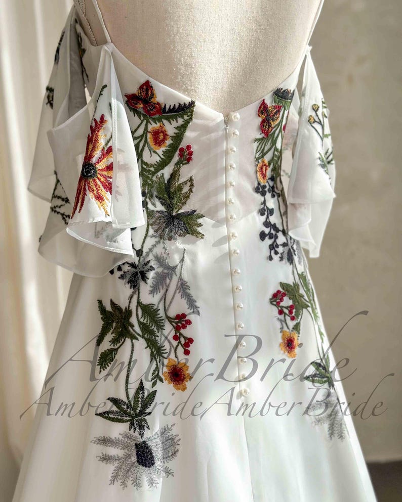 May include: White wedding dress with floral embroidery in red, yellow, green, and black. The dress features a button-down back and off-the-shoulder sleeves with ruffled detailing. The dress has a flowing, elegant design.