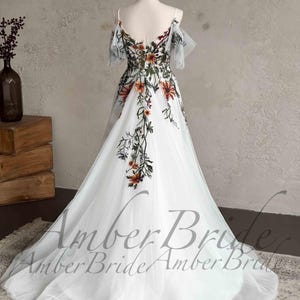 May include: A white tulle gown with a floral embroidery design. The gown has a sweetheart neckline and off-the-shoulder straps. The embroidery features a variety of flowers in shades of orange, red, and green.