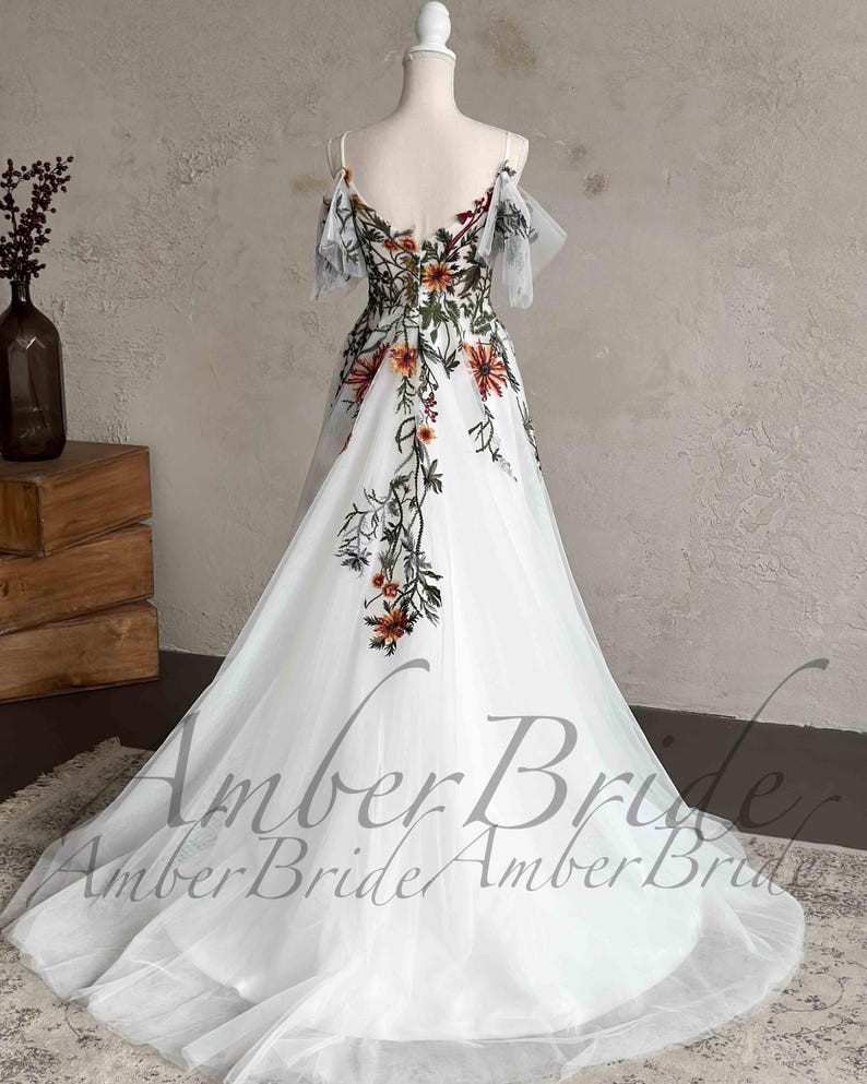 May include: A white tulle gown with a floral embroidery design. The gown has a sweetheart neckline and off-the-shoulder straps. The embroidery features a variety of flowers in shades of orange, red, and green.